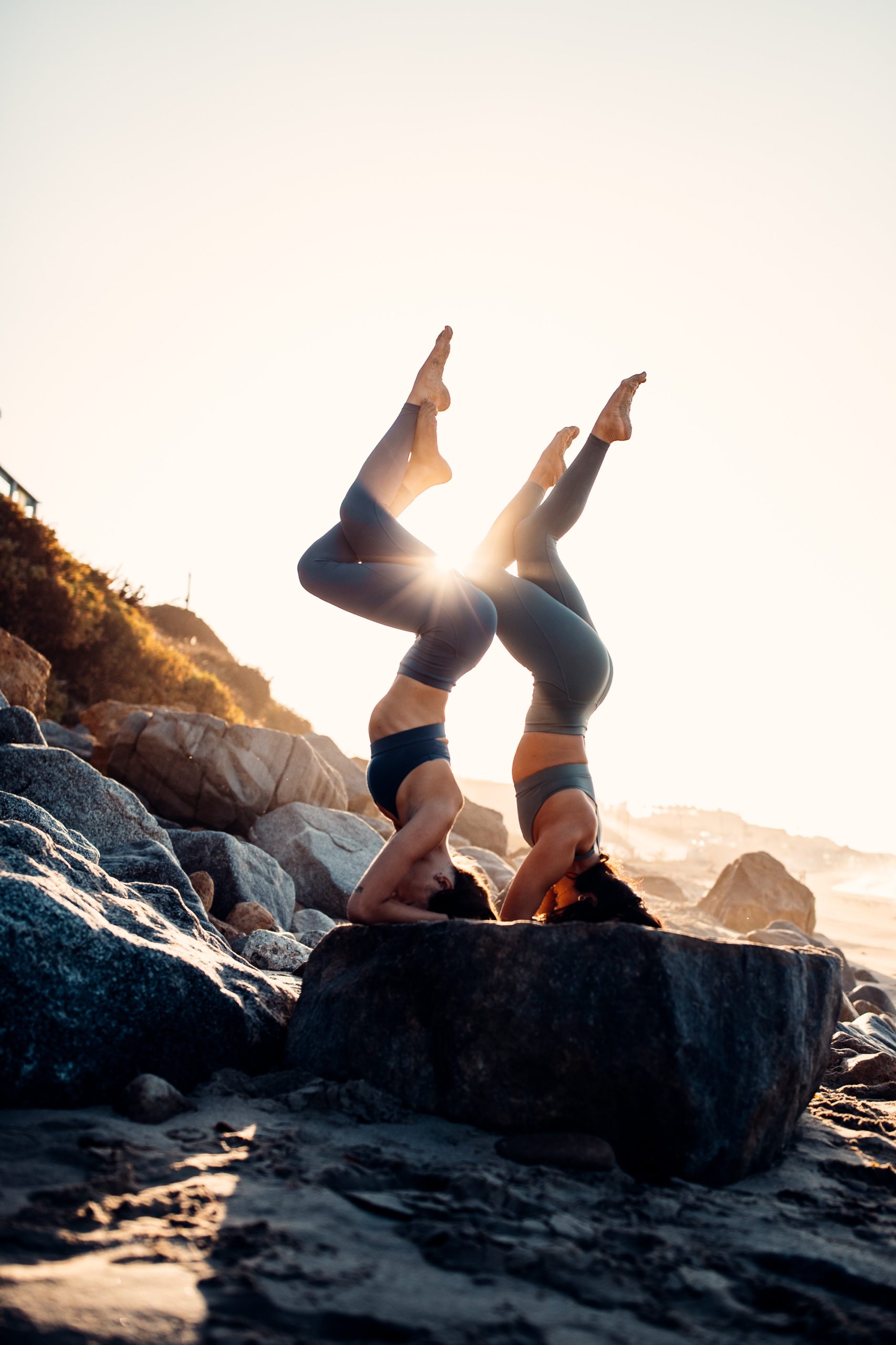 2 girls doing headstand on top of a stone by the beach