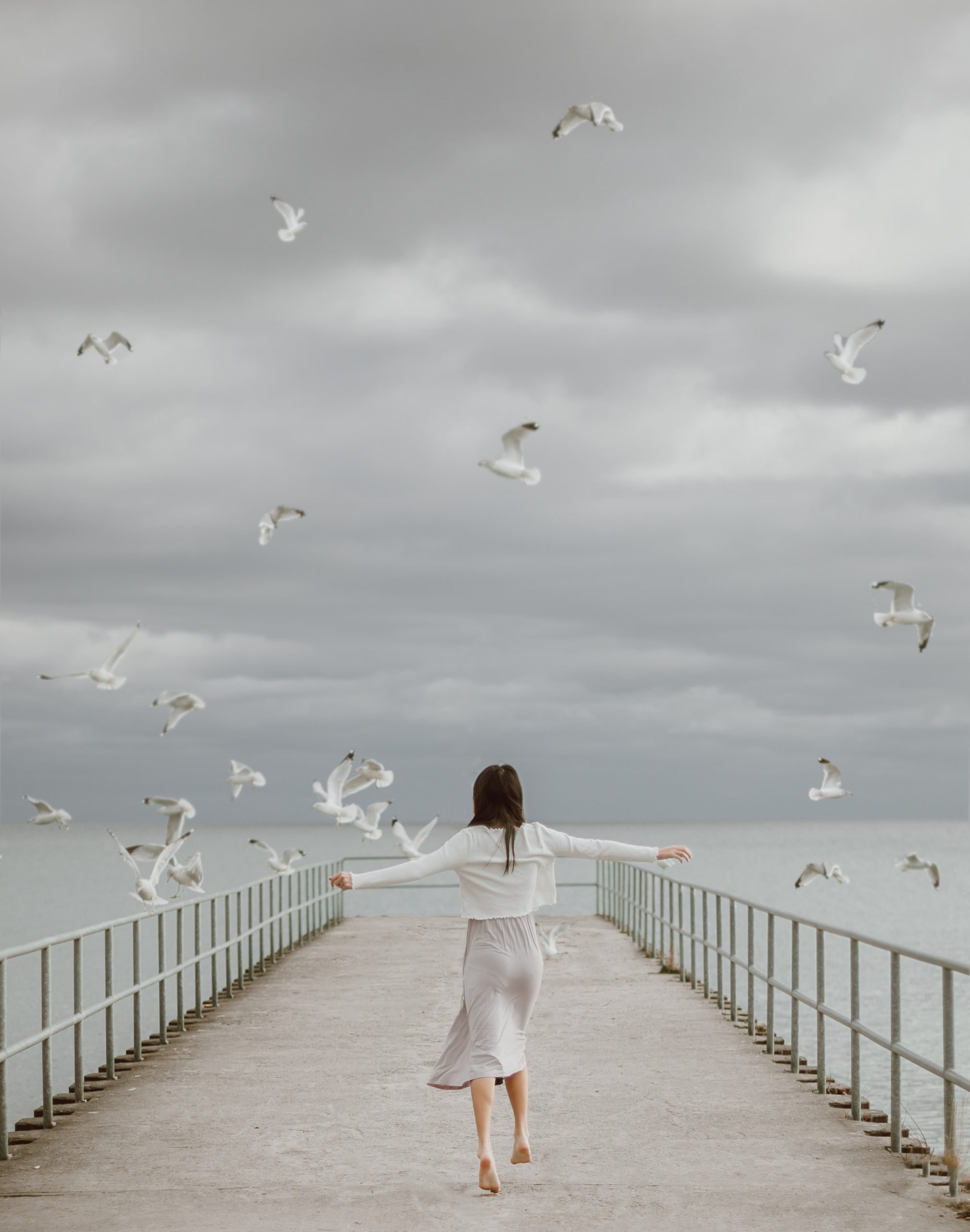 Girl on the dock spreading her arms. and birds are flying around her.