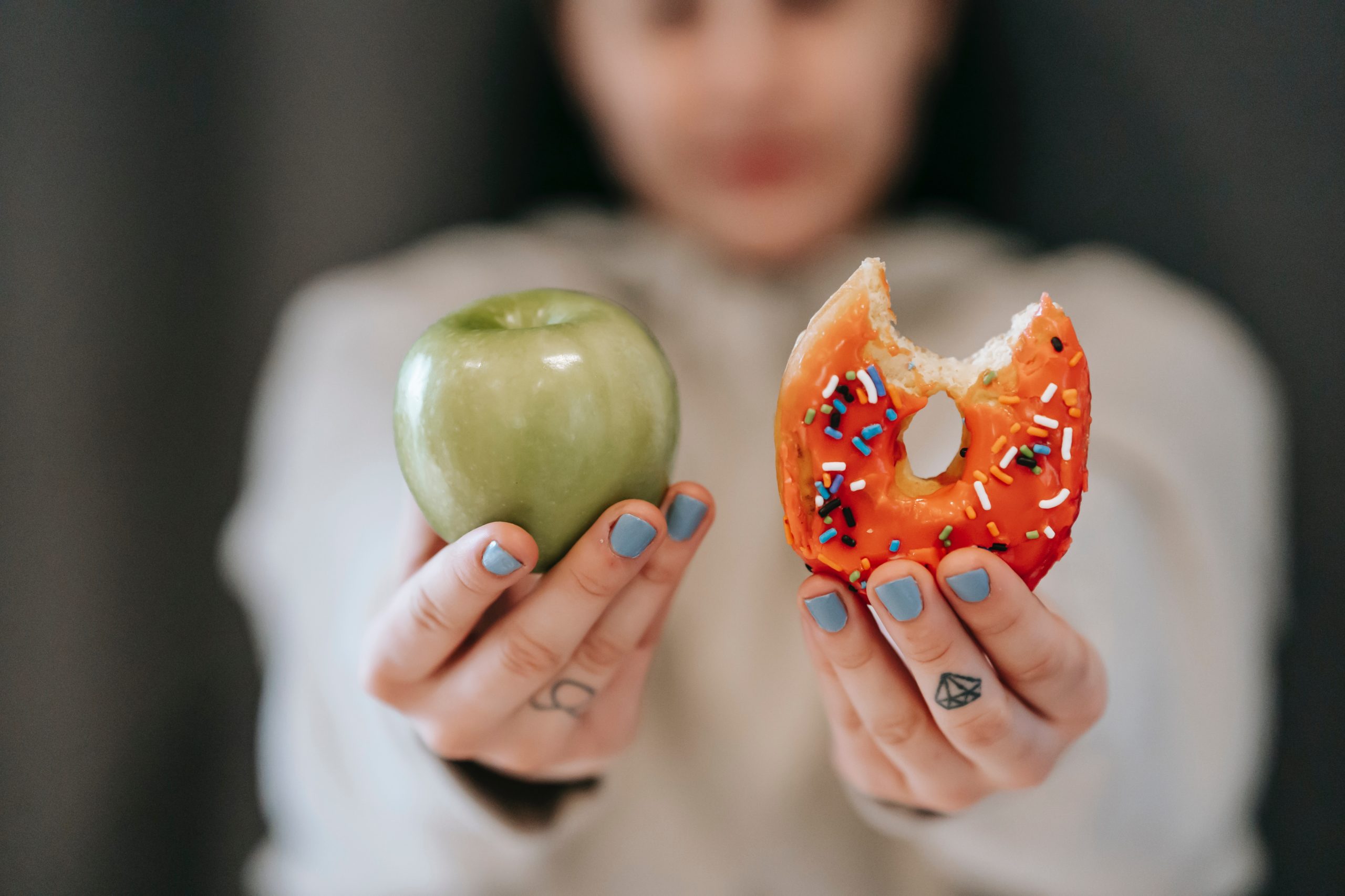 Woman holding 2 objects. Right hand is apple. Left hand is doughnut.